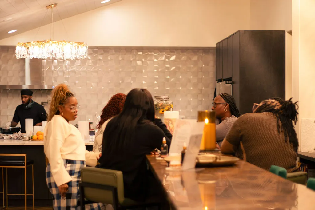 Women seated along a marble counter in conversation while a chef cooks behind them