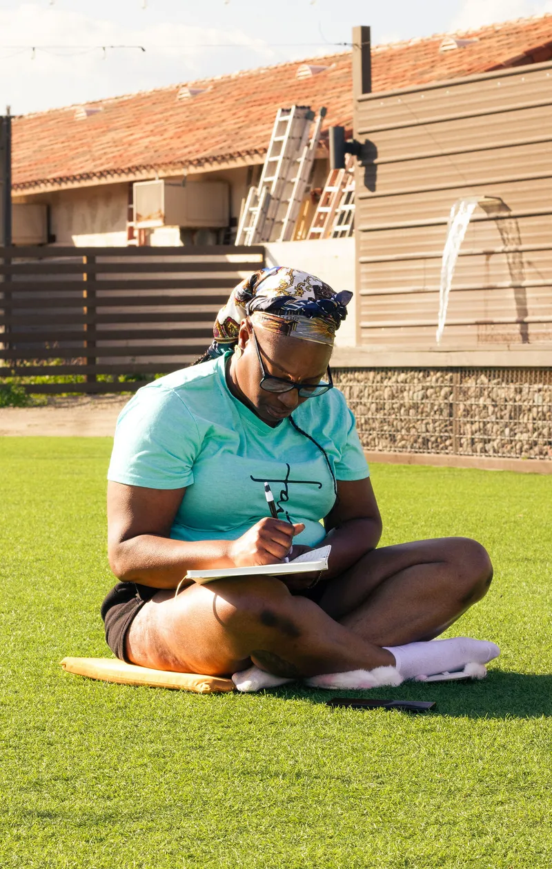 A woman in a headwrap writing in a notebook on the grass in golden light