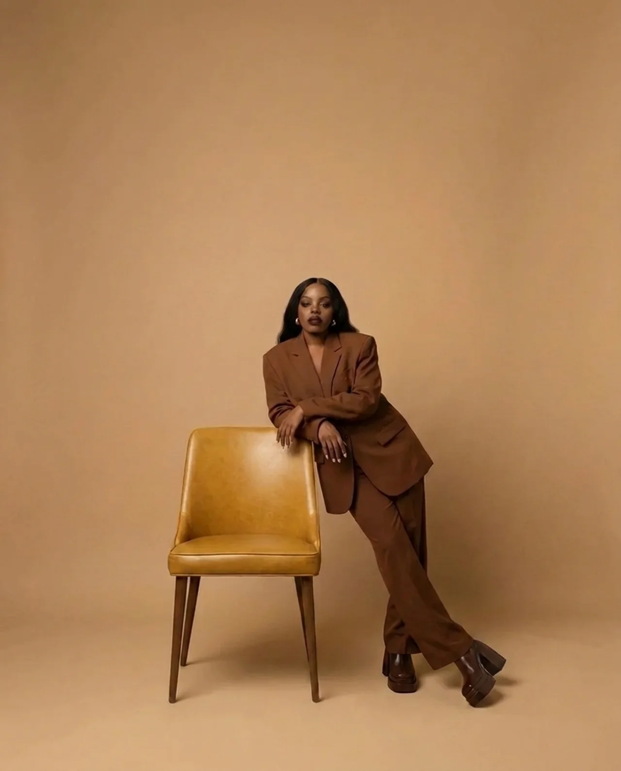 A woman in a warm brown suit leaning beside a brass chair, inviting you to take your seat.