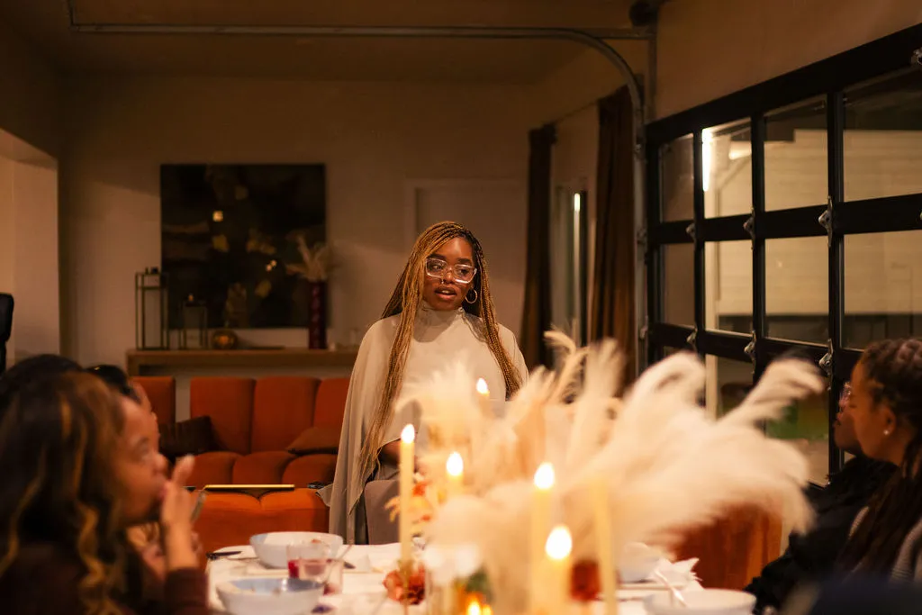 A woman speaking at a candlelit Solumere gathering, surrounded by pampas grass and seated guests
